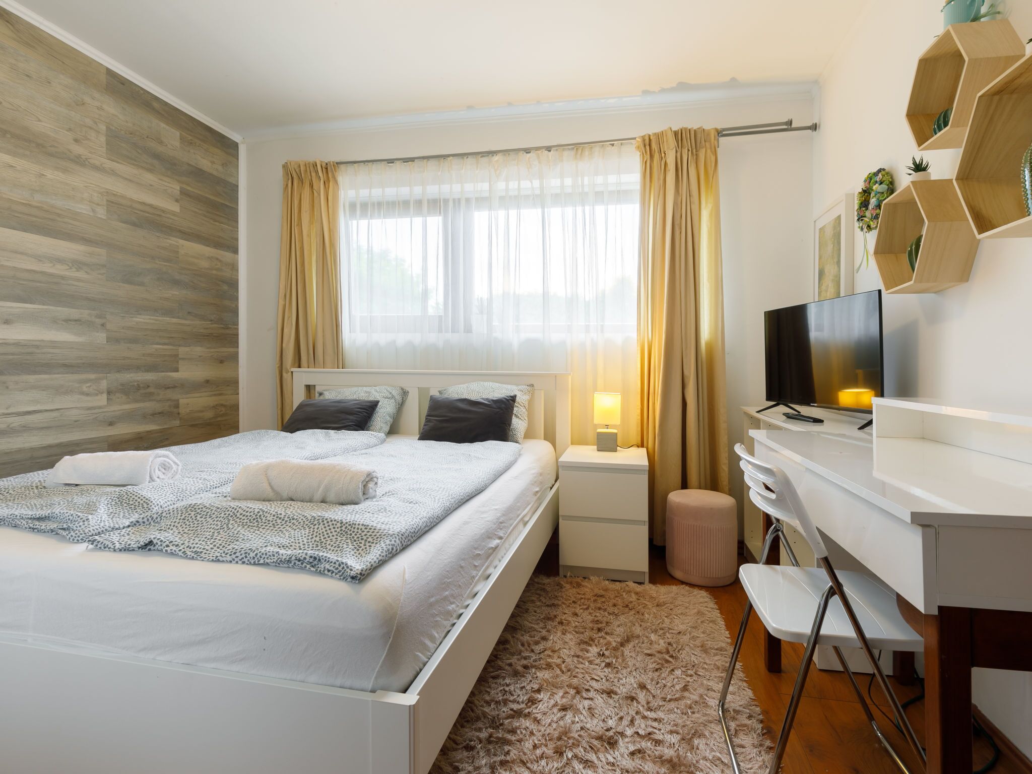 Une chambre à coucher moderne, avec un lit, des tables de chevet, un bureau, une chaise, un téléviseur et une fenêtre avec des rideaux.