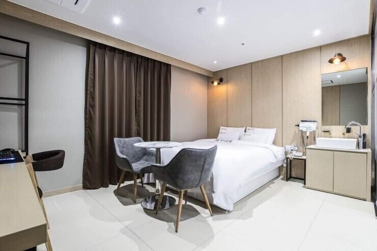 A modern hotel room with a bed, bedside table, chair, desk, and a bathroom with a sink and mirror.