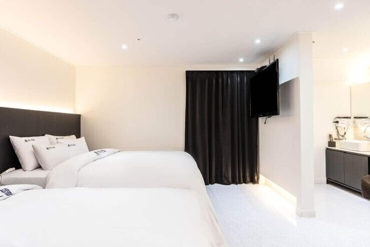 A modern hotel room with a large bed, a flat-screen TV mounted on the wall, a dark curtain, and a bathroom with a white sink and mirror.
