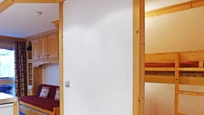 A compact room with bunk beds, a small kitchenette, and a single bed with a red blanket.