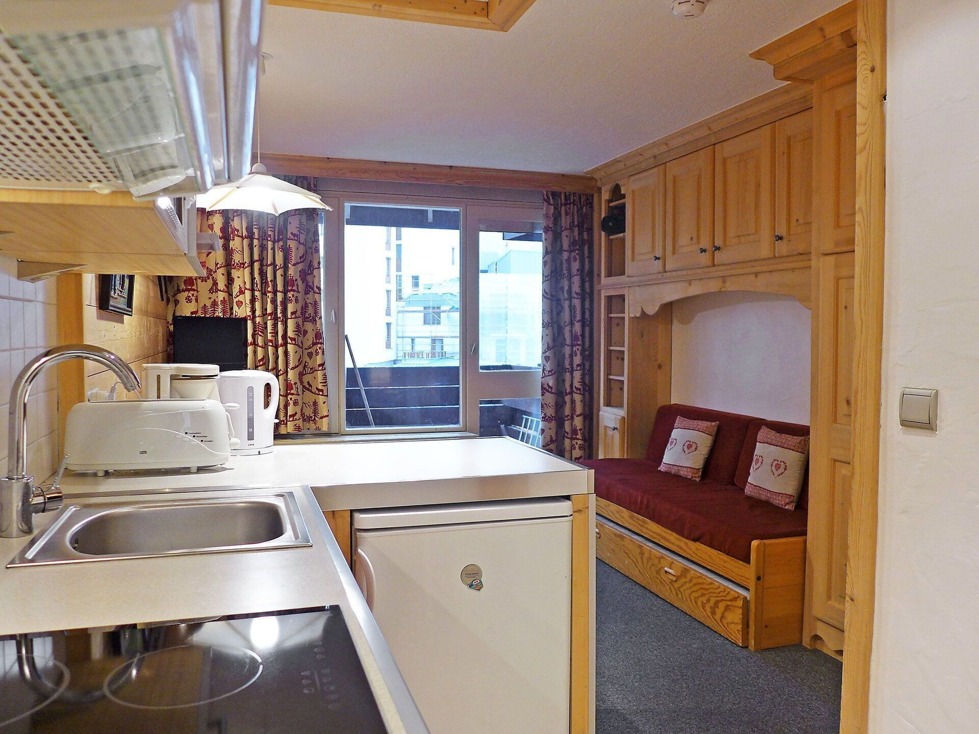 A compact kitchen with a sink, toaster, and refrigerator, a window with curtains, and a wooden bunk bed with a red mattress.