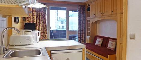 A compact kitchen with a sink, toaster, and refrigerator, a window with curtains, and a wooden bunk bed with a red mattress.