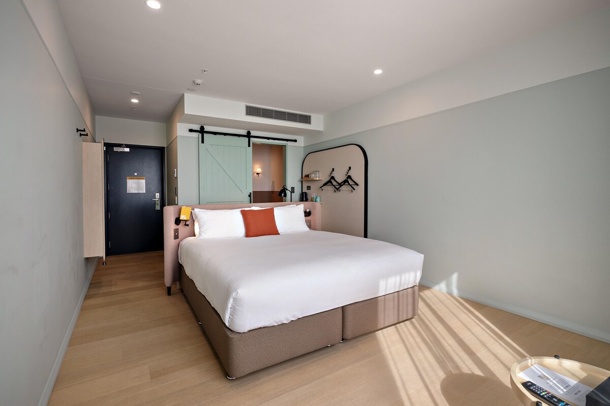 A modern hotel room with a large bed, a desk with a lamp, a wardrobe, and a view of the hallway.