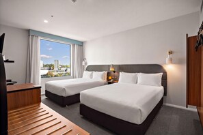 A hotel room with two beds, a desk, a chair, a view of the city, and a large window with curtains.
