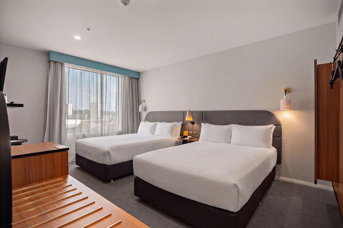 A hotel room with two beds, a desk, a chair, a TV, and a window with curtains.