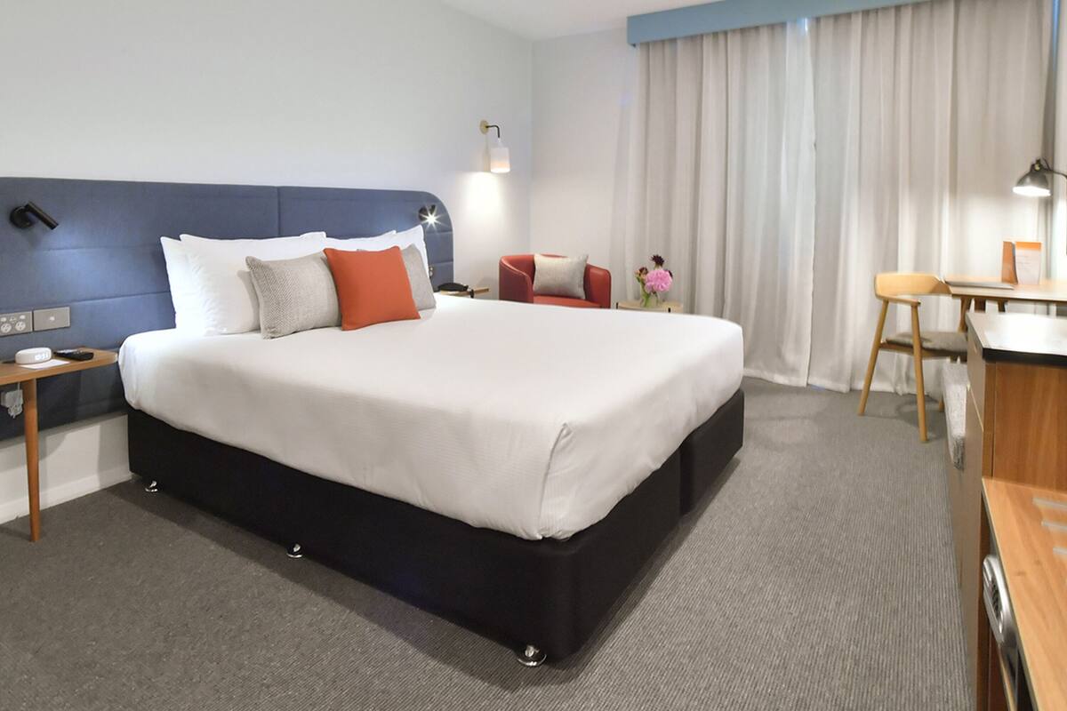 A hotel room with a large bed, a desk, a chair, a nightstand, and a lamp.