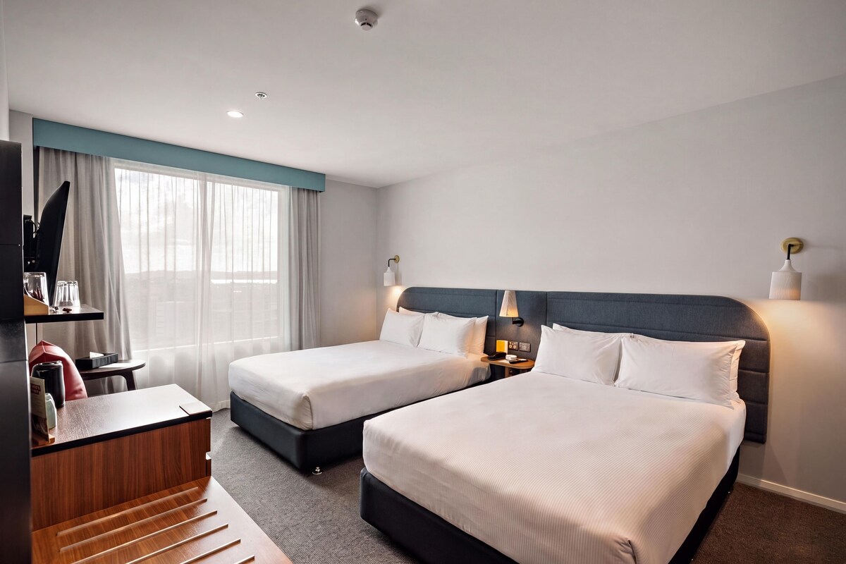 A hotel room with two beds, a desk, a chair, a TV, and a window with curtains.