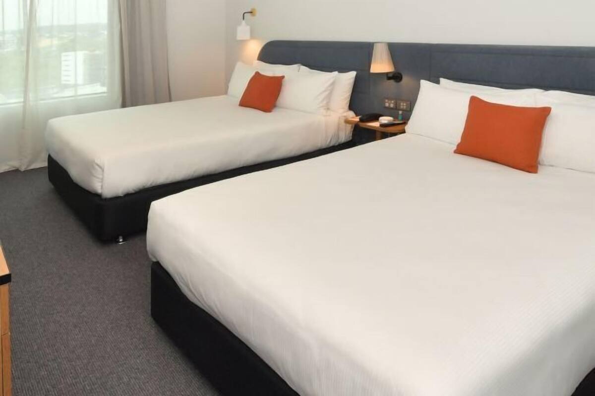 A hotel room with two beds, a nightstand, a lamp, and a window with curtains.