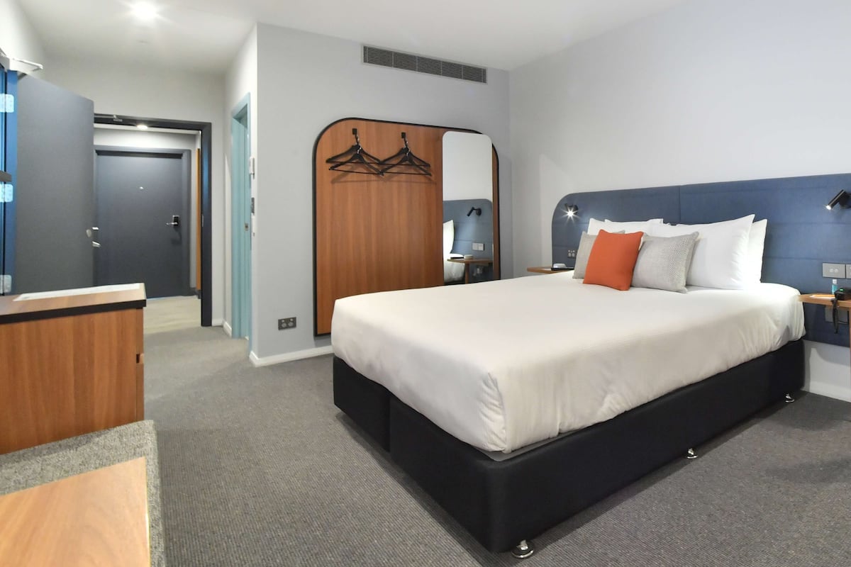A modern hotel room with a large bed, a wooden wardrobe, a mirror, and a desk.