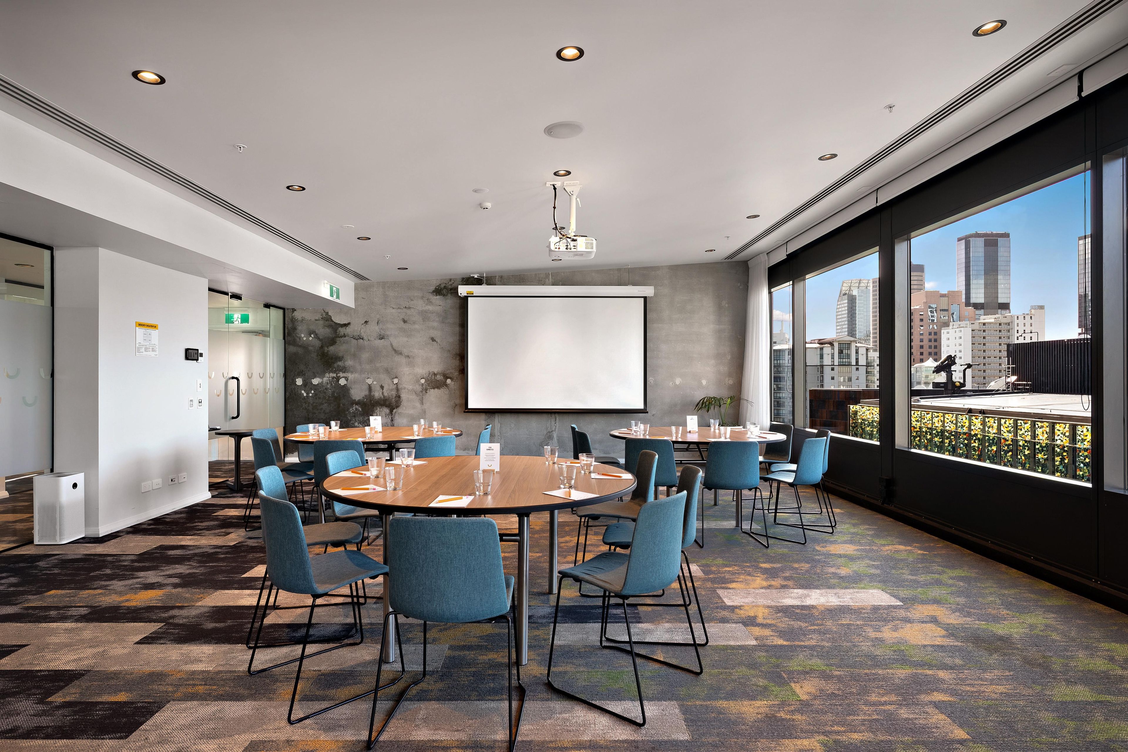 A modern conference room with round tables, blue chairs, a projector screen, and large windows offering a city view.