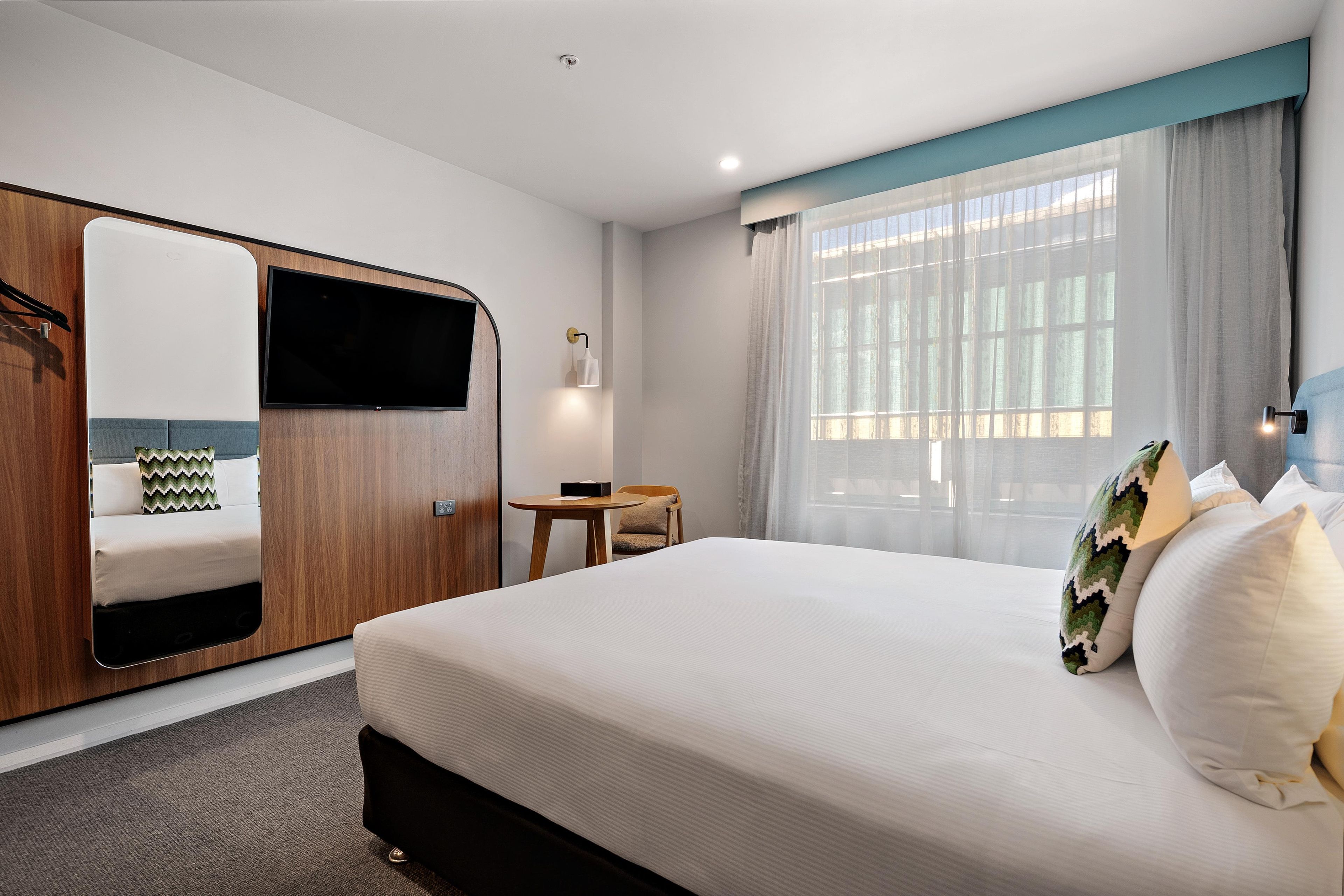 Habitación de hotel con cama, escritorio, silla, televisor y ventana con cortinas.