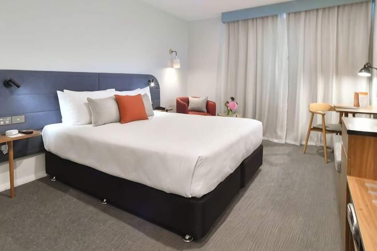 A hotel room with a large bed, a desk, a chair, a nightstand, and a window with curtains.