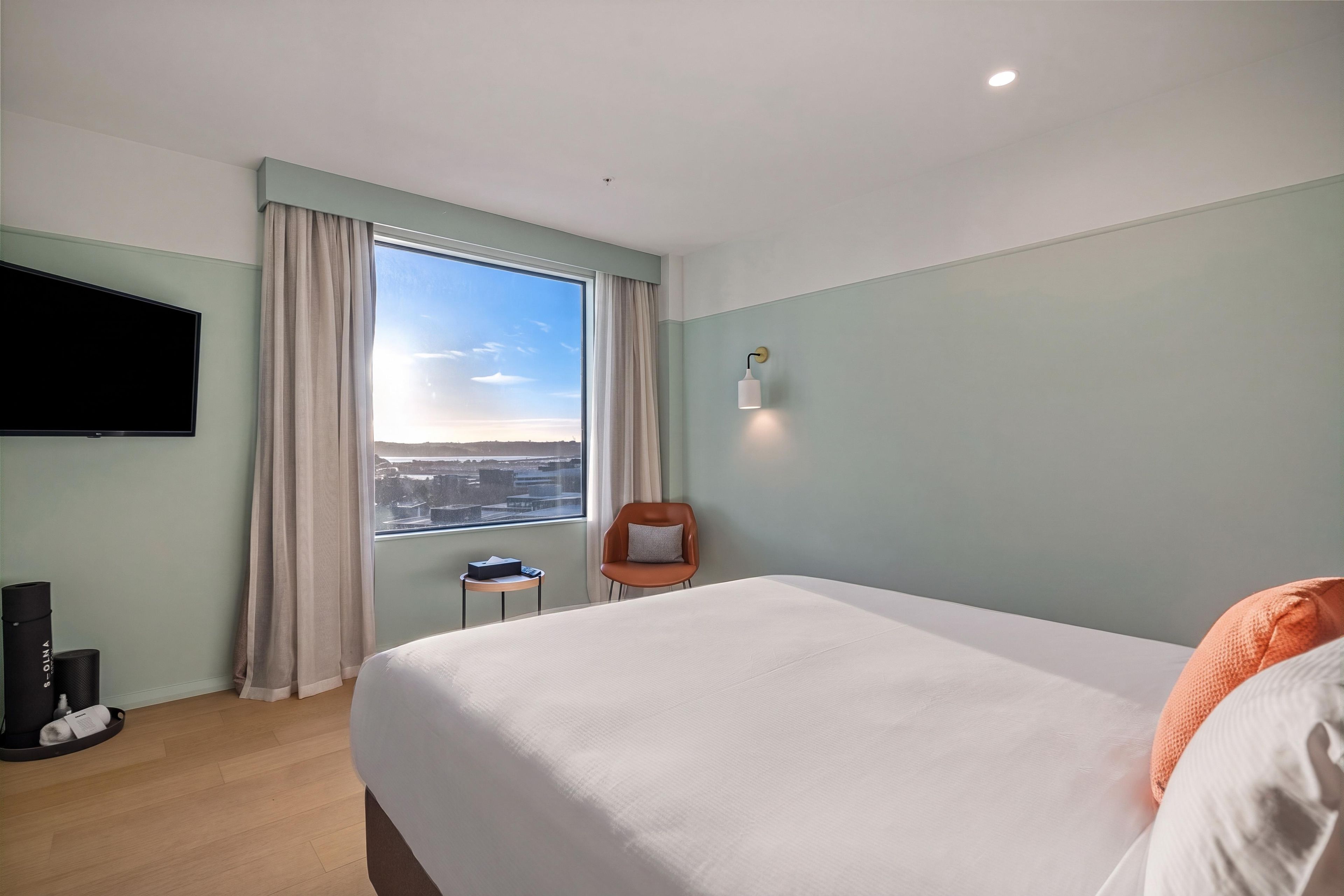 Habitación de hotel con una cama grande, televisor montado en la pared, una silla y ventana con vistas a la ciudad.