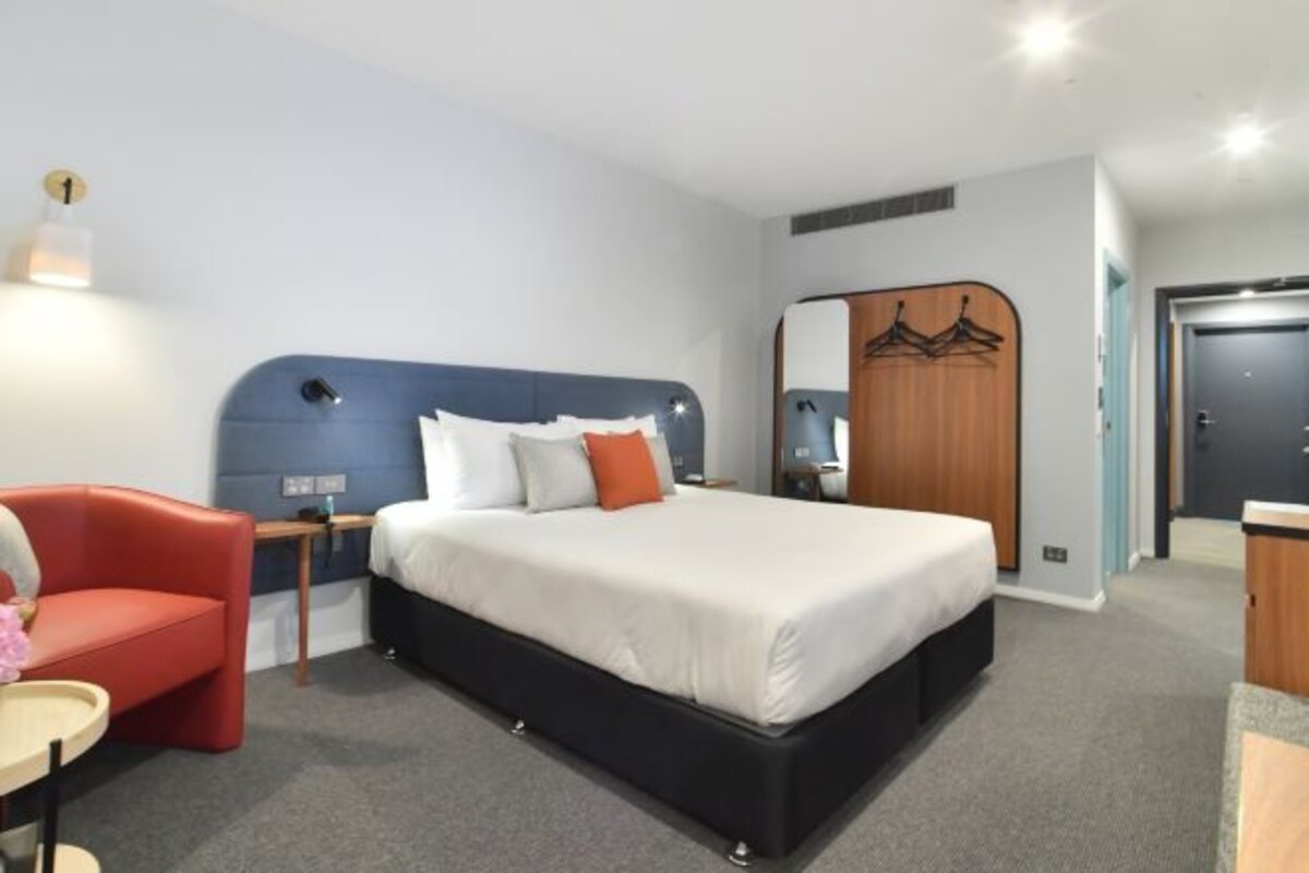 A hotel room with a bed, a red chair, a small table, a mirror, and a wardrobe.