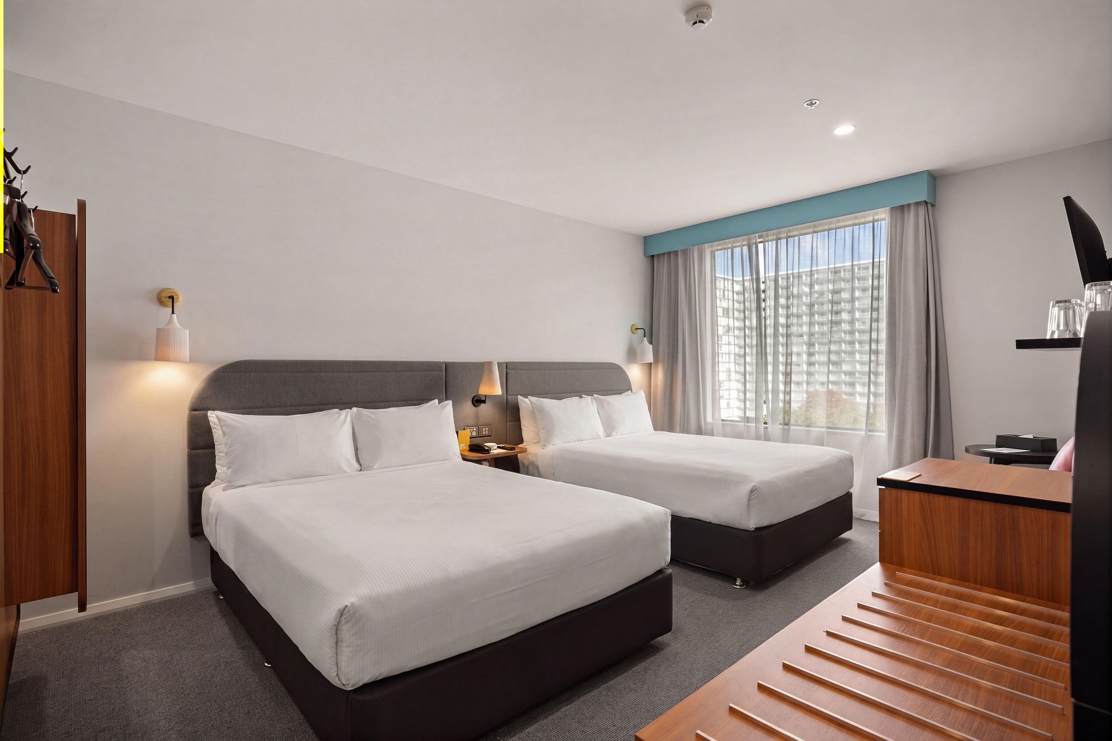 Quarto de hotel com duas camas, uma escrivaninha, uma cadeira e janela com cortinas.