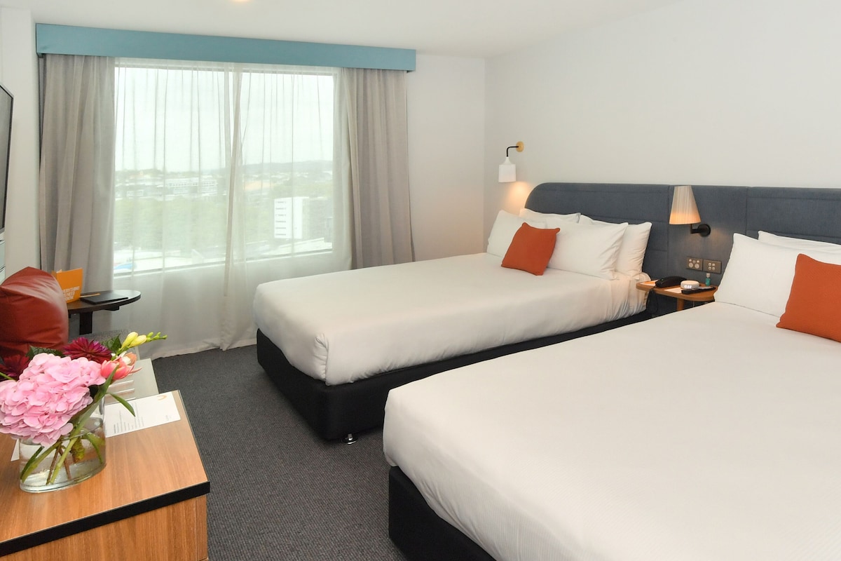 A hotel room with two beds, a desk with a vase of flowers, a nightstand with a lamp, and a view of the city through large windows.
