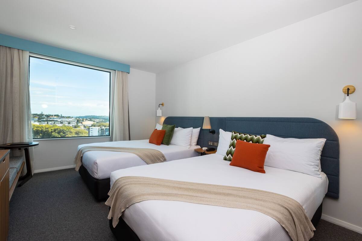 A hotel room with two beds, a desk, a chair, a window with curtains, and a view of the city.