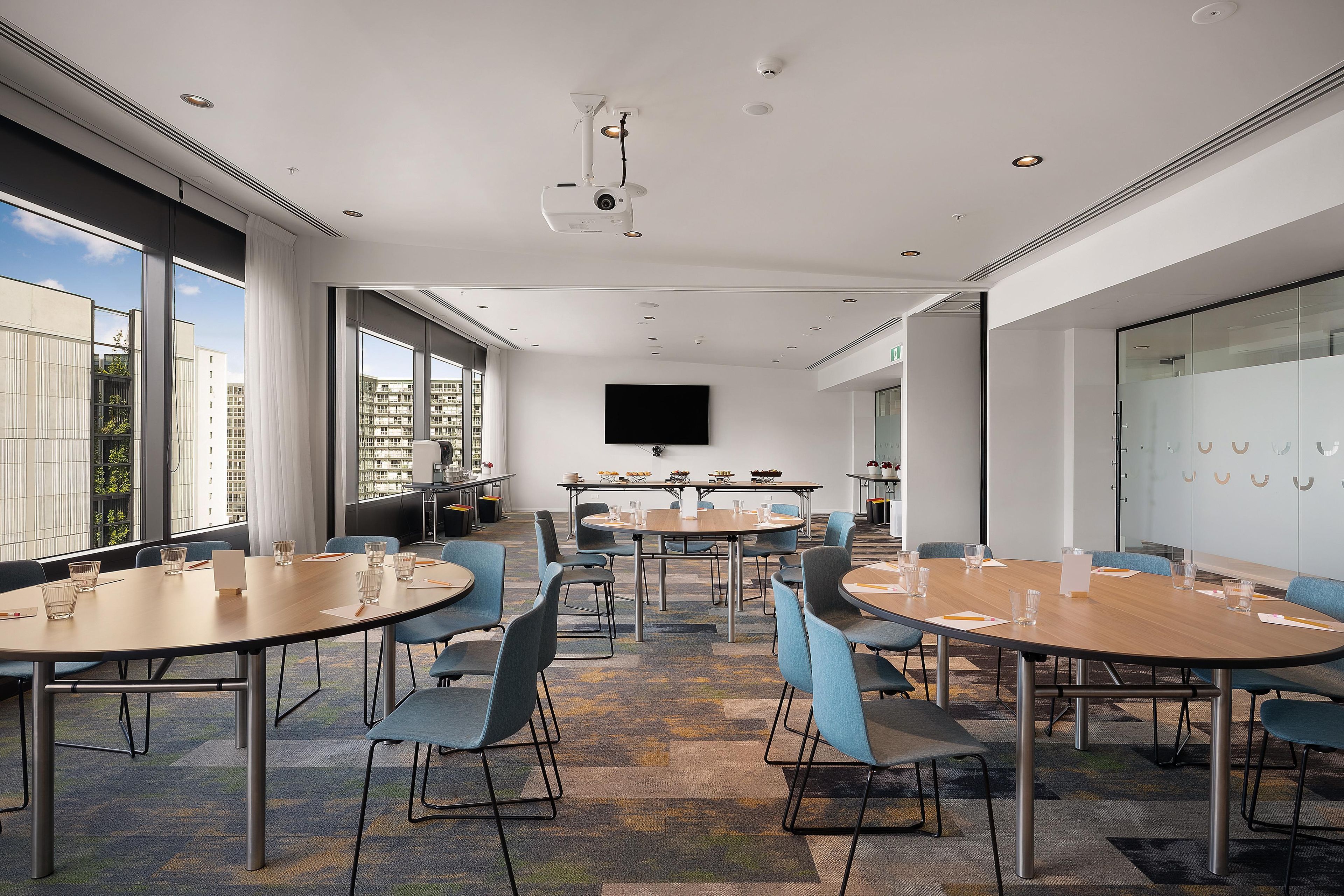 Un salón de conferencias con varias mesas y sillas, una pantalla grande y vistas a edificios a través de amplias ventanas.