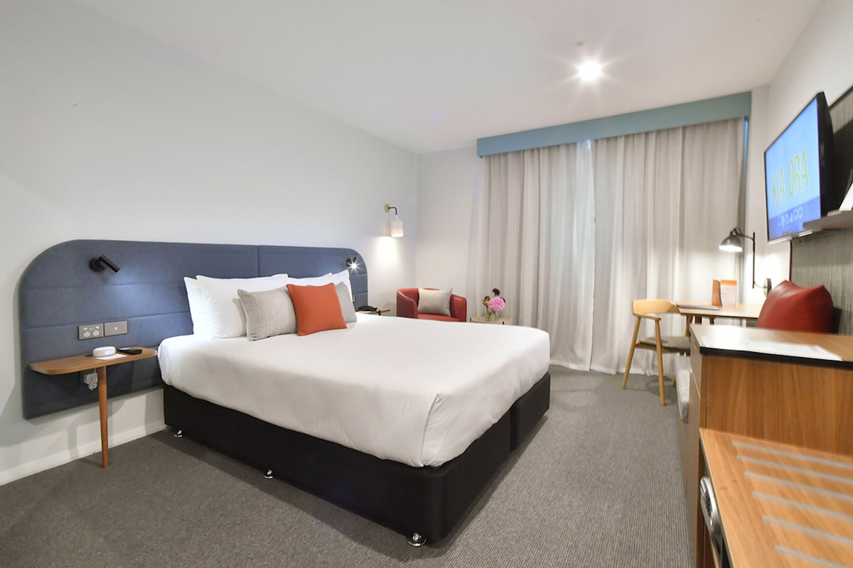 A hotel room with a large bed, a desk, a chair, a TV, and a window with curtains.