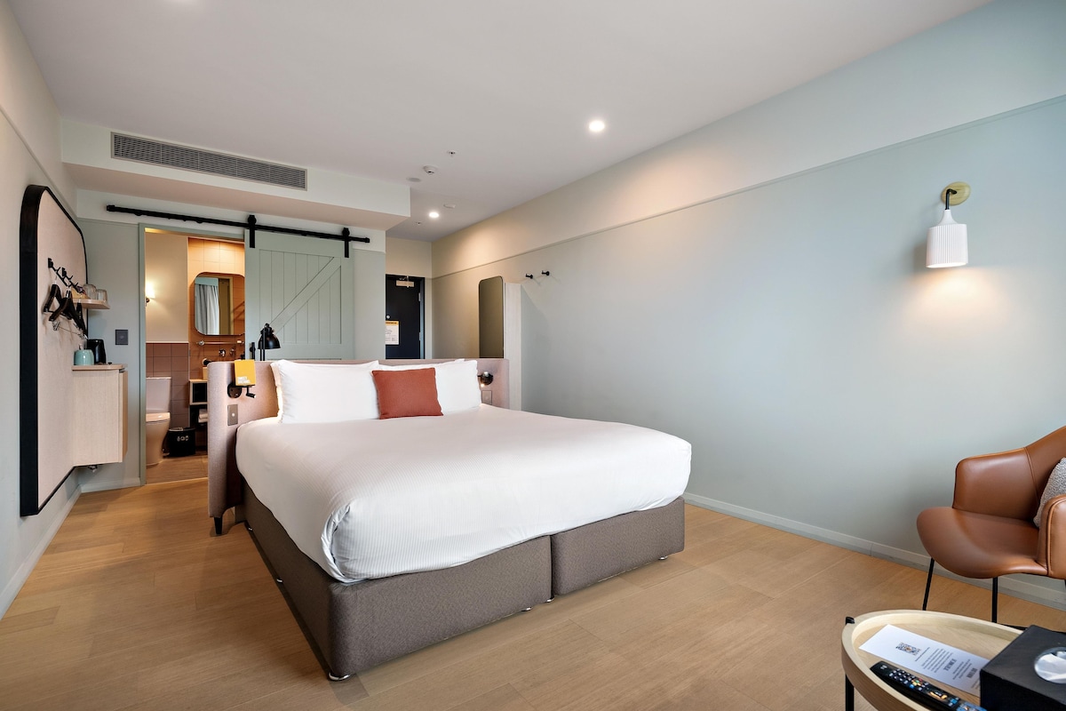 A modern hotel room with a large bed, a desk, a chair, and a bathroom visible through an open door.