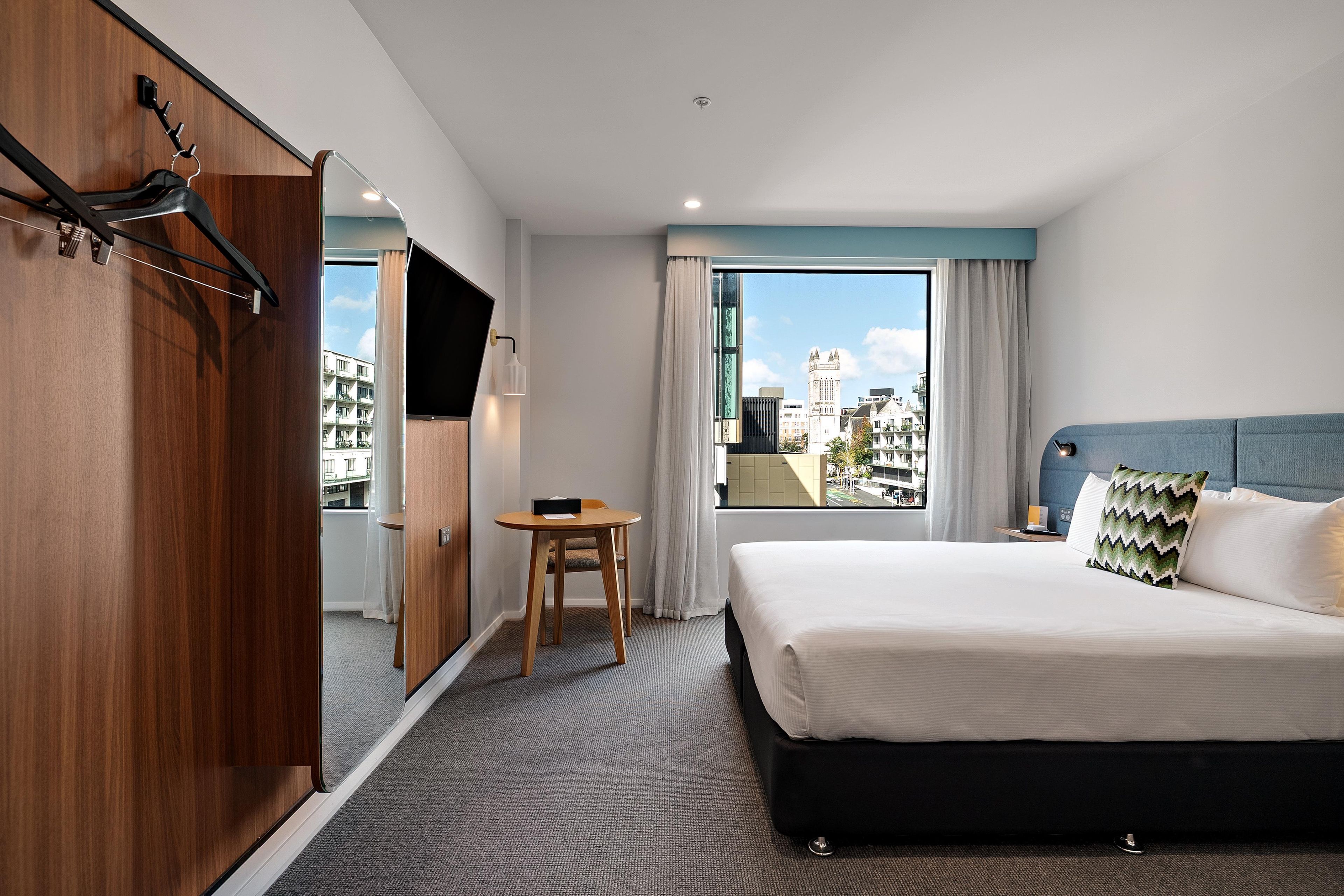 A hotel room with a bed, a desk, a chair, a wardrobe, and a window with a city view.