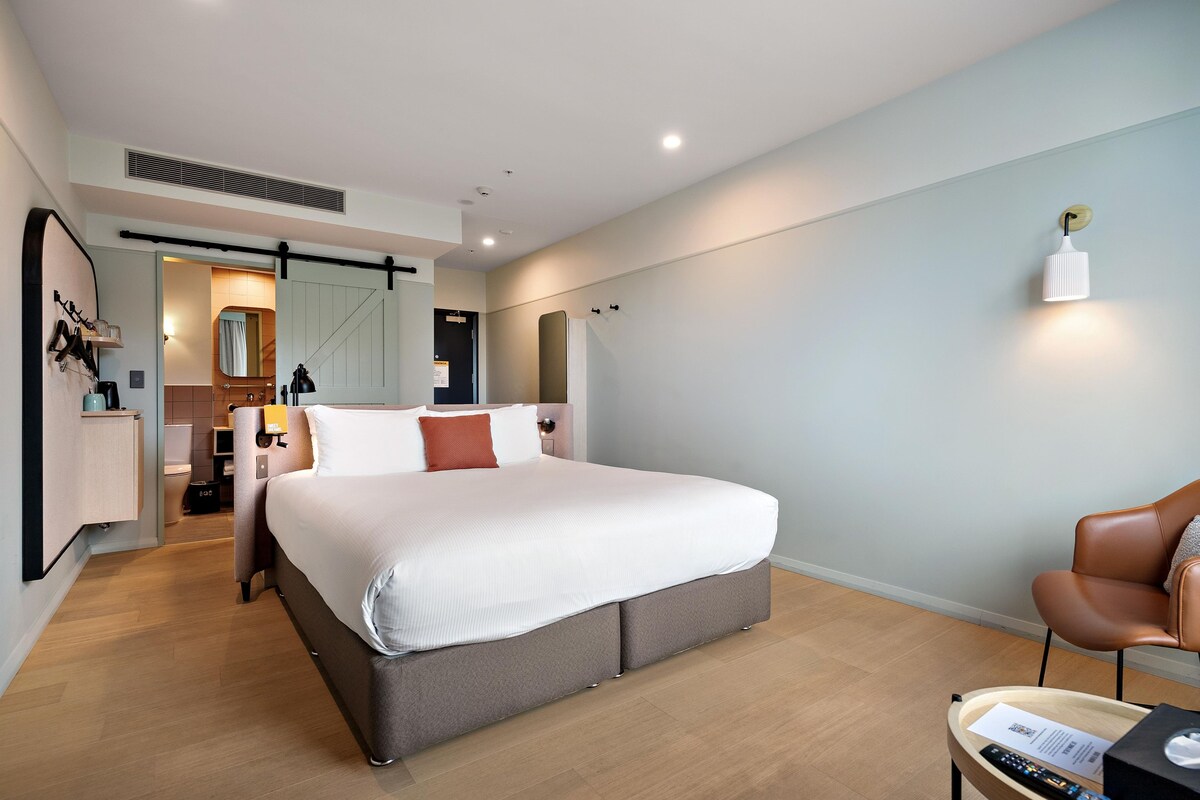 A modern hotel room with a large bed, a desk, a chair, and a bathroom visible through an open door.