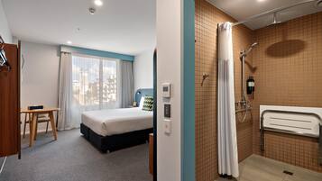 Una habitación de hotel con cama, escritorio, ventana con cortinas, baño con ducha y pared con azulejos.