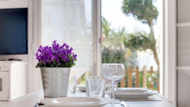 Uma mesa de jantar com toalha branca, um vaso com planta de flores roxas, copos transparentes e um prato branco.