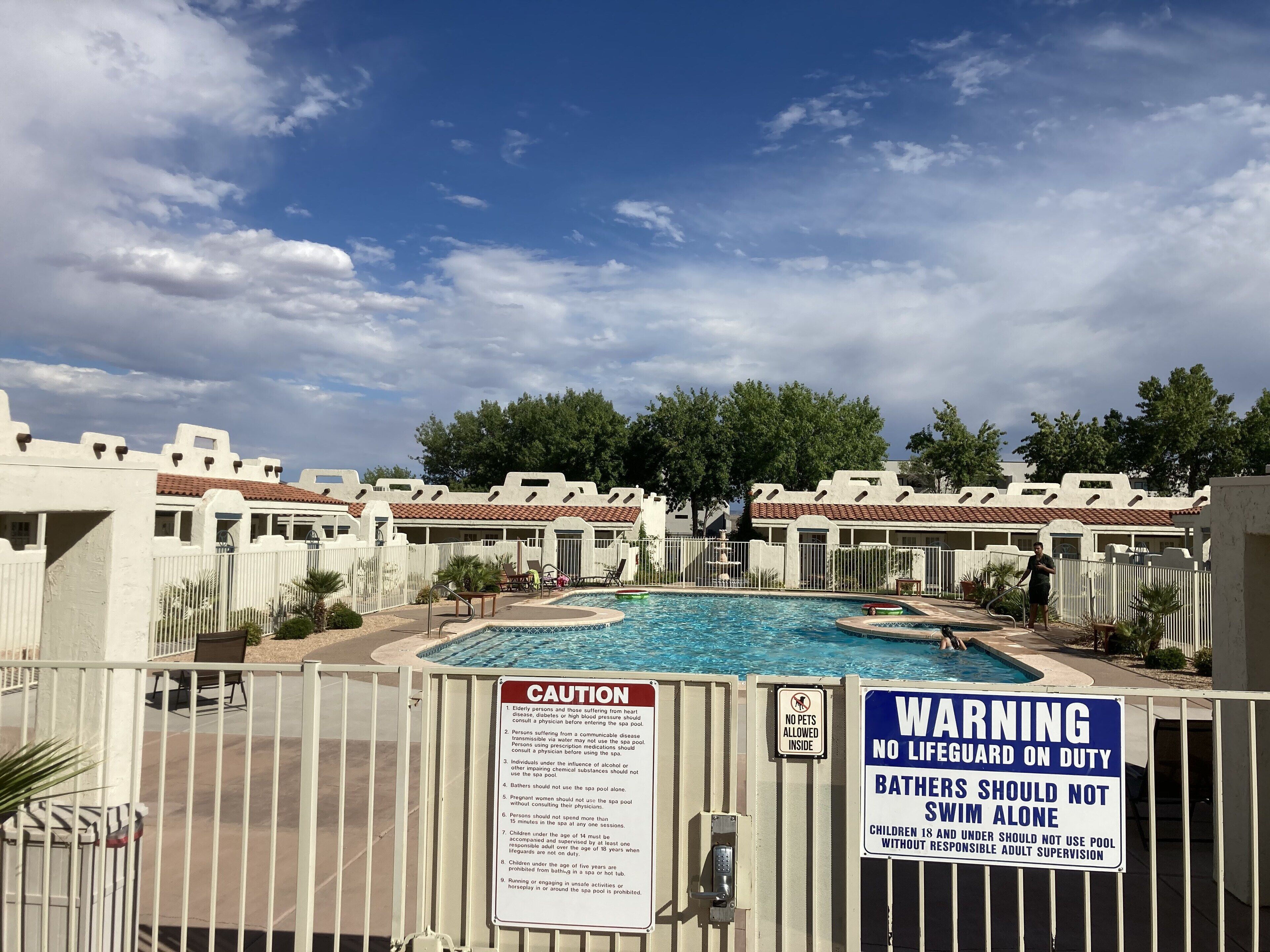 Zona de piscina con varias piscinas, rodeada de edificios blancos con techos de tejas rojas. Hay carteles que indican que no se permiten mascotas y que no hay socorrista de servicio.