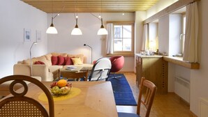 A cozy living room with a wooden ceiling, a sofa, a dining table with chairs, and a kitchen area with a window.