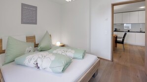 Un dormitorio con cama, almohadas, una mesita de noche, un aplique de pared y una cocina visible a través de una puerta abierta.