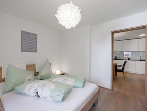 A bedroom with a bed, pillows, a bedside table, a wall-mounted light fixture, and a kitchen visible through an open door.