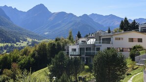 Un edificio moderno con fachada de piedra, rodeado de abundante vegetación y montañas al fondo.