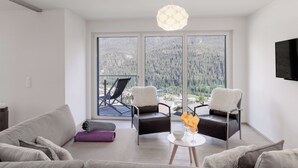Una sala de estar moderna con un sofá, dos sillones, una mesita con un tazón y vasos, y una lámpara colgante. Amplias ventanas ofrecen vistas a un área boscosa.