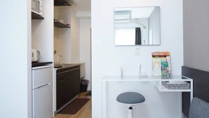 Uma cozinha compacta com geladeira, fogão e micro-ondas. Um banquinho pequeno e uma prateleira com livros também são visíveis.