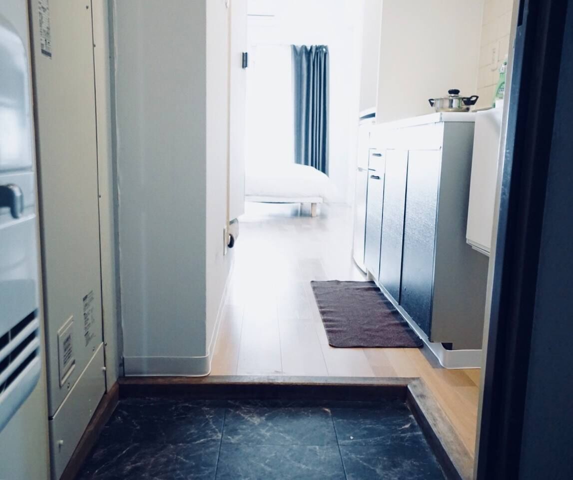 A narrow hallway with a wooden floor leading to a bedroom with a bed and a window.