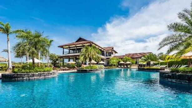 A large swimming pool with a modern resort building in the background, surrounded by palm trees and lounge areas.