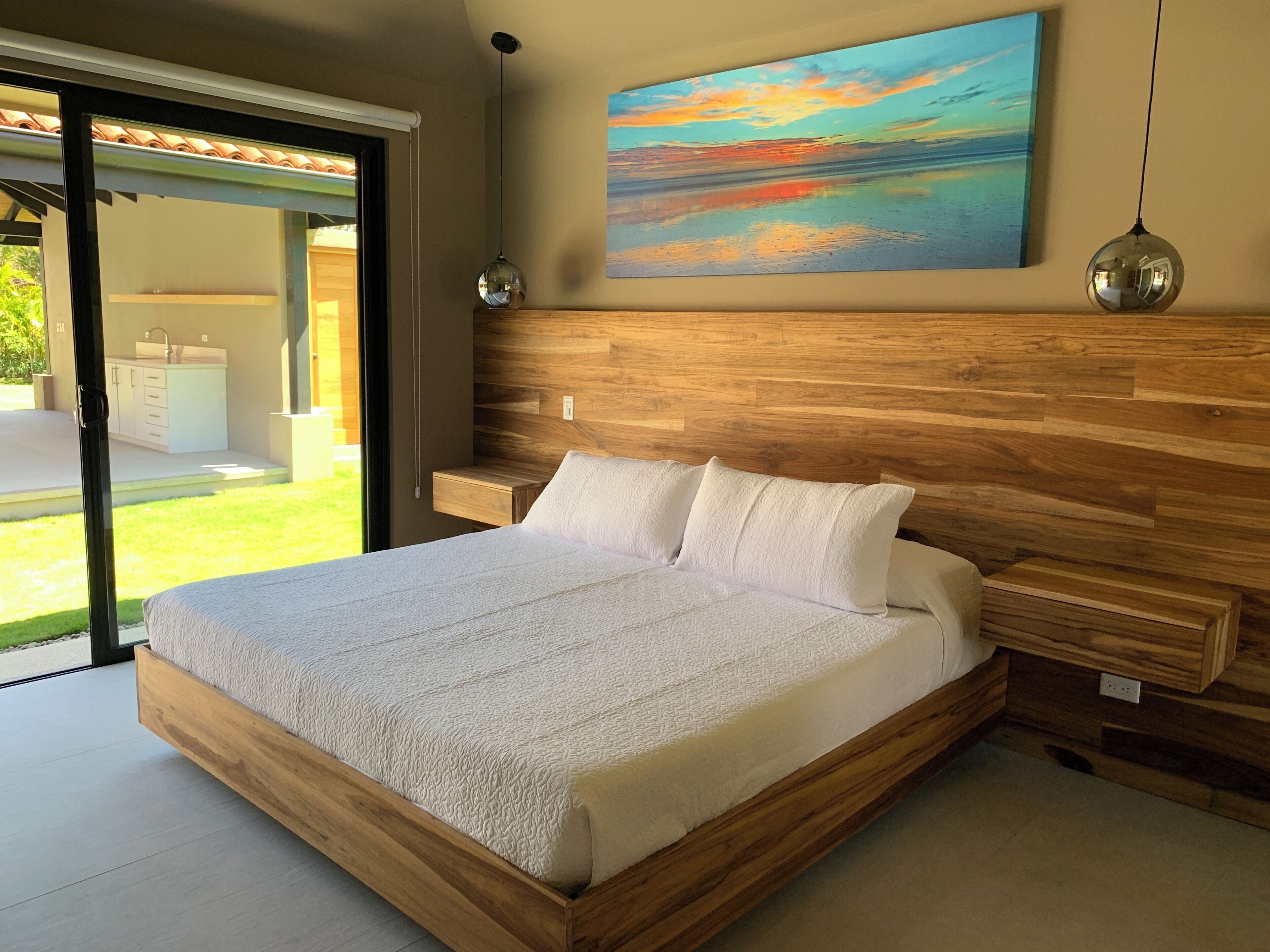 A modern bedroom with a wooden headboard, a large bed with white bedding, a painting of a sunset, and a view of a garden through a sliding glass door.
