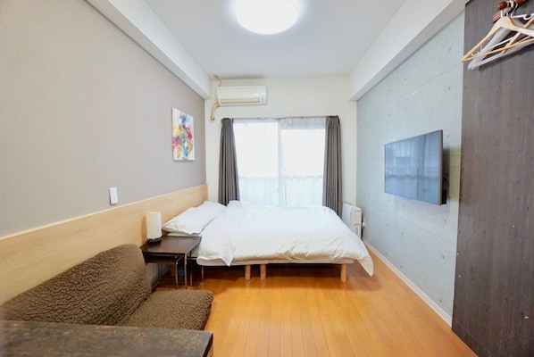 A hotel room with a bed, a small table, a chair, a sofa, an air conditioning unit, a TV, and a window with curtains.