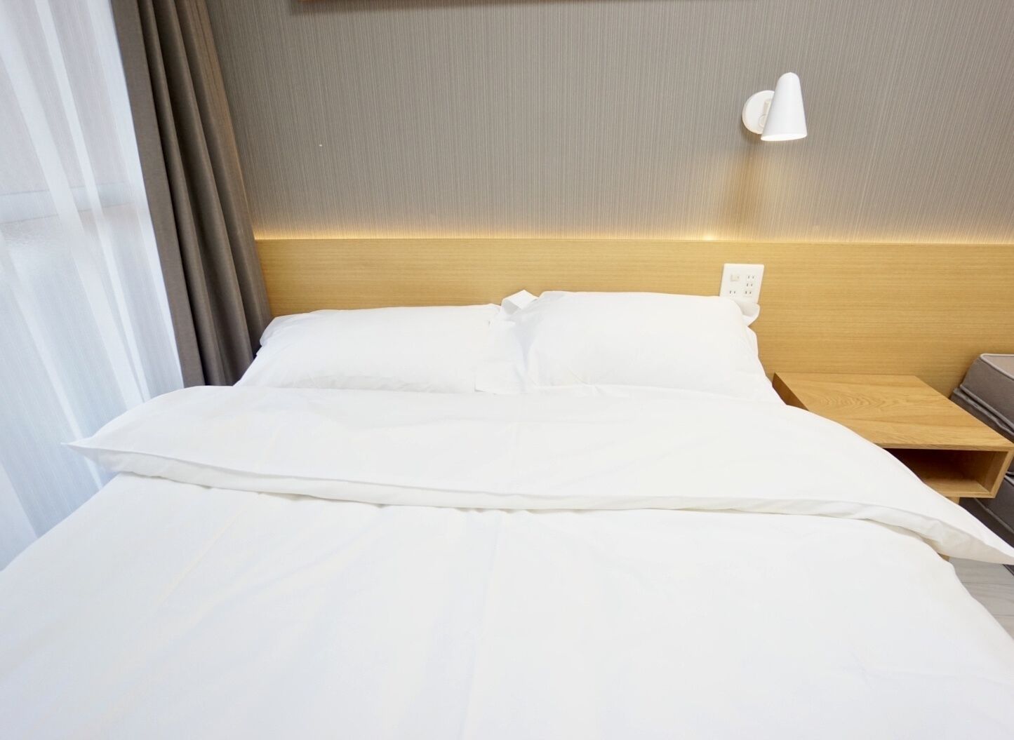 Habitación de hotel con una cama bien hecha, cabecera de madera, mesita de noche y una lámpara fijada a la pared.
