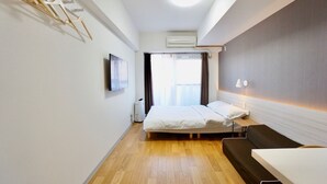 A modern hotel room with a bed, a bedside table, a desk, and a chair. There is a window with curtains, an air conditioning unit, and a wall-mounted TV. - Hostel 758 Nagoya 1K (Nagoya)