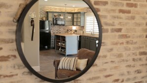 A circular mirror reflecting a kitchen with a brick wall, a microwave, a sink, and a brown leather chair with a patterned cushion.