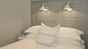 A neatly made bed with white linens and striped pillows, two wall-mounted lamps, and a small bedside table with a plant.