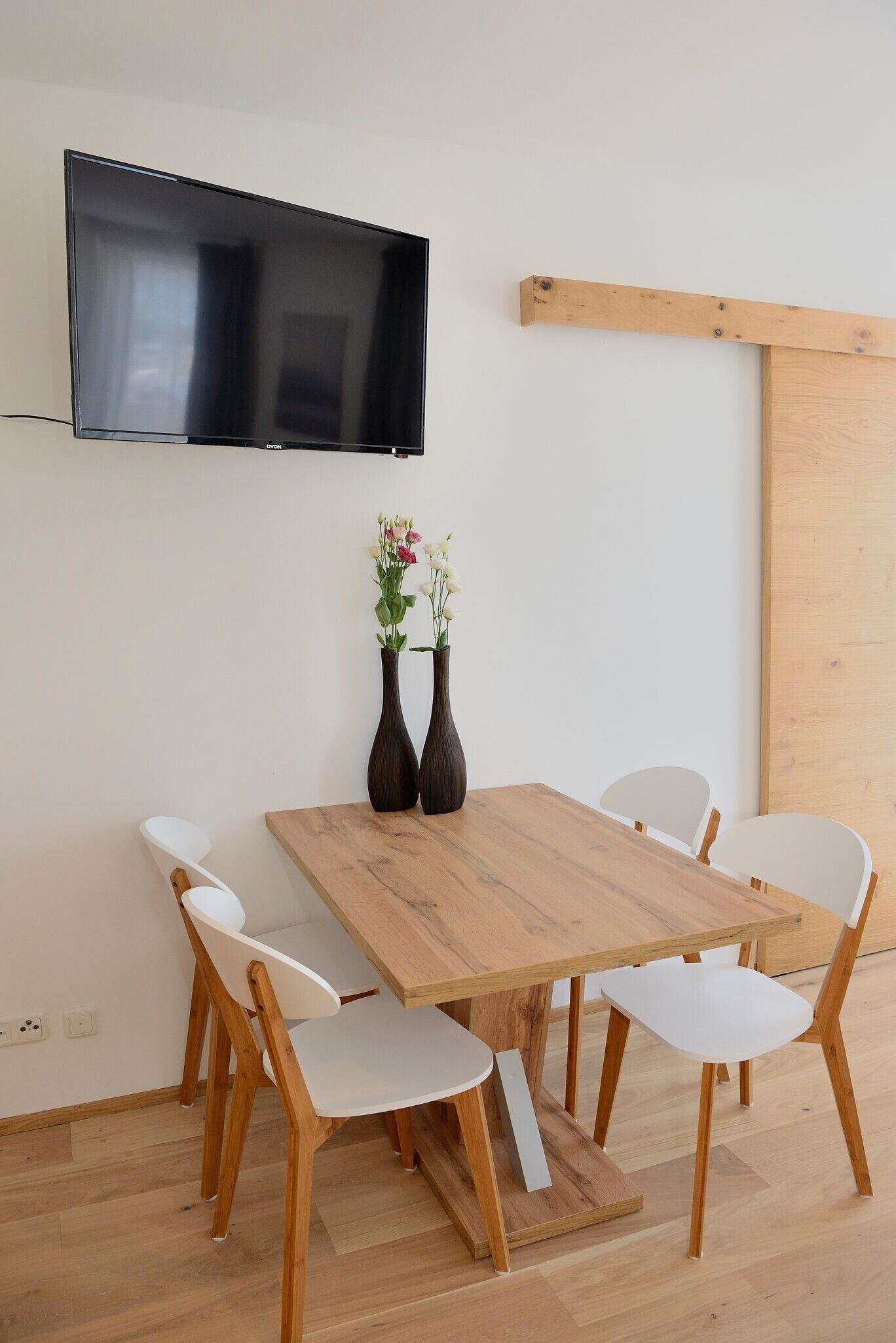 A dining area with a wooden table, white chairs, two vases with flowers, and a mounted TV.