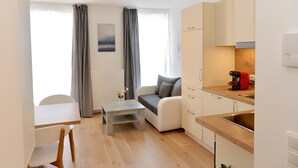 Ein modernes Wohnzimmer mit einer Couch, einem Couchtisch, einem Esstisch mit Stühlen, einer Küchenzeile mit Spüle und Schränken sowie einem Fenster mit Vorhängen.