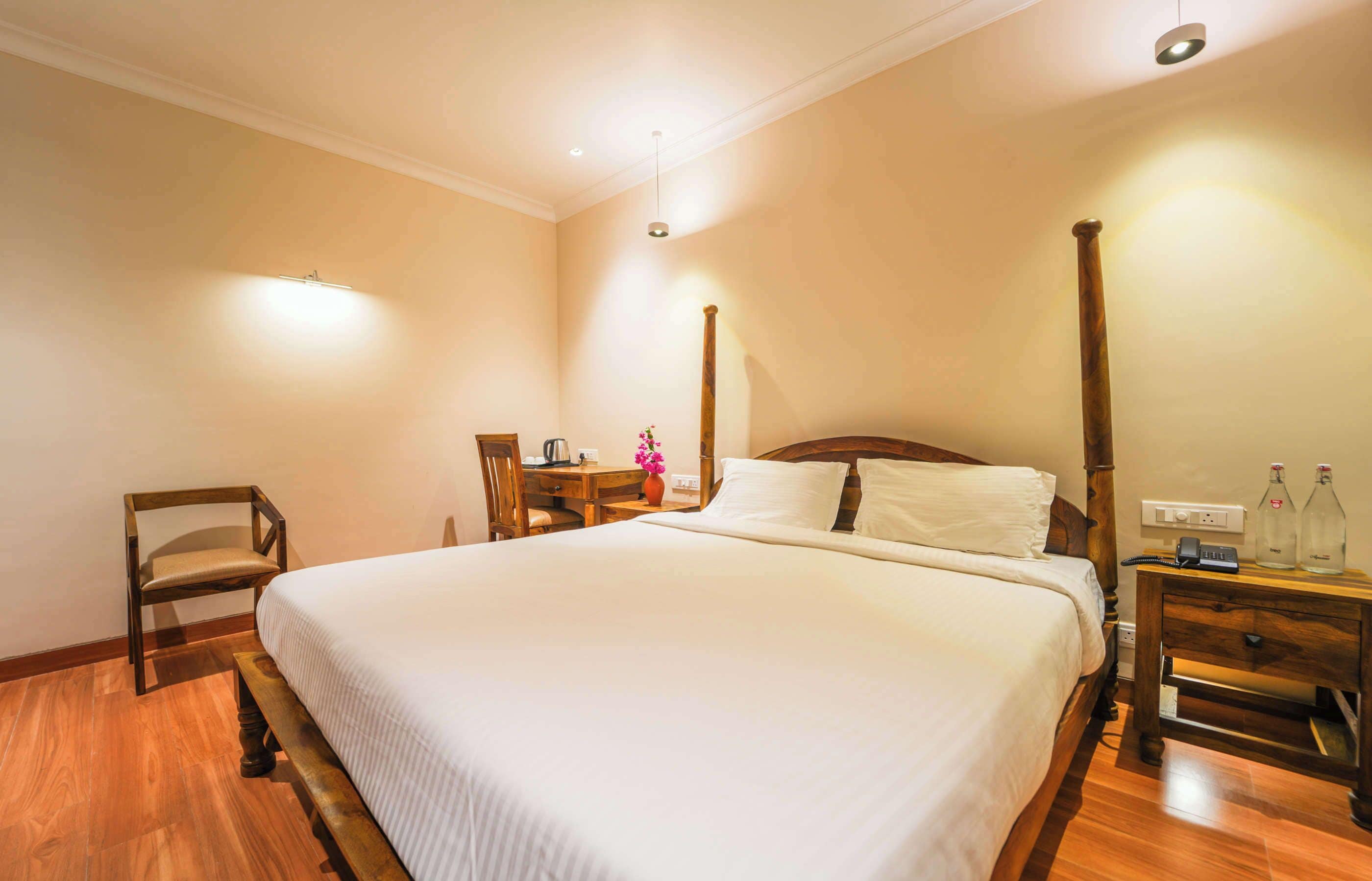 A neatly arranged hotel room with a large bed, a wooden chair, a desk with a lamp, and a nightstand with bottles.
