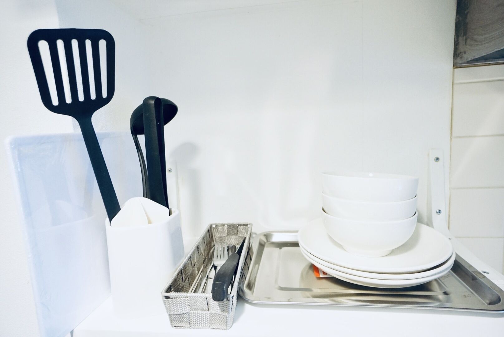 A kitchen area with a white countertop, a metal tray, a basket with utensils, and a stack of white bowls.
