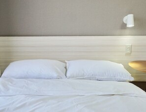 Una cama bien tendida con sábanas blancas, una lámpara fijada a la pared y un enchufe en la pared.
