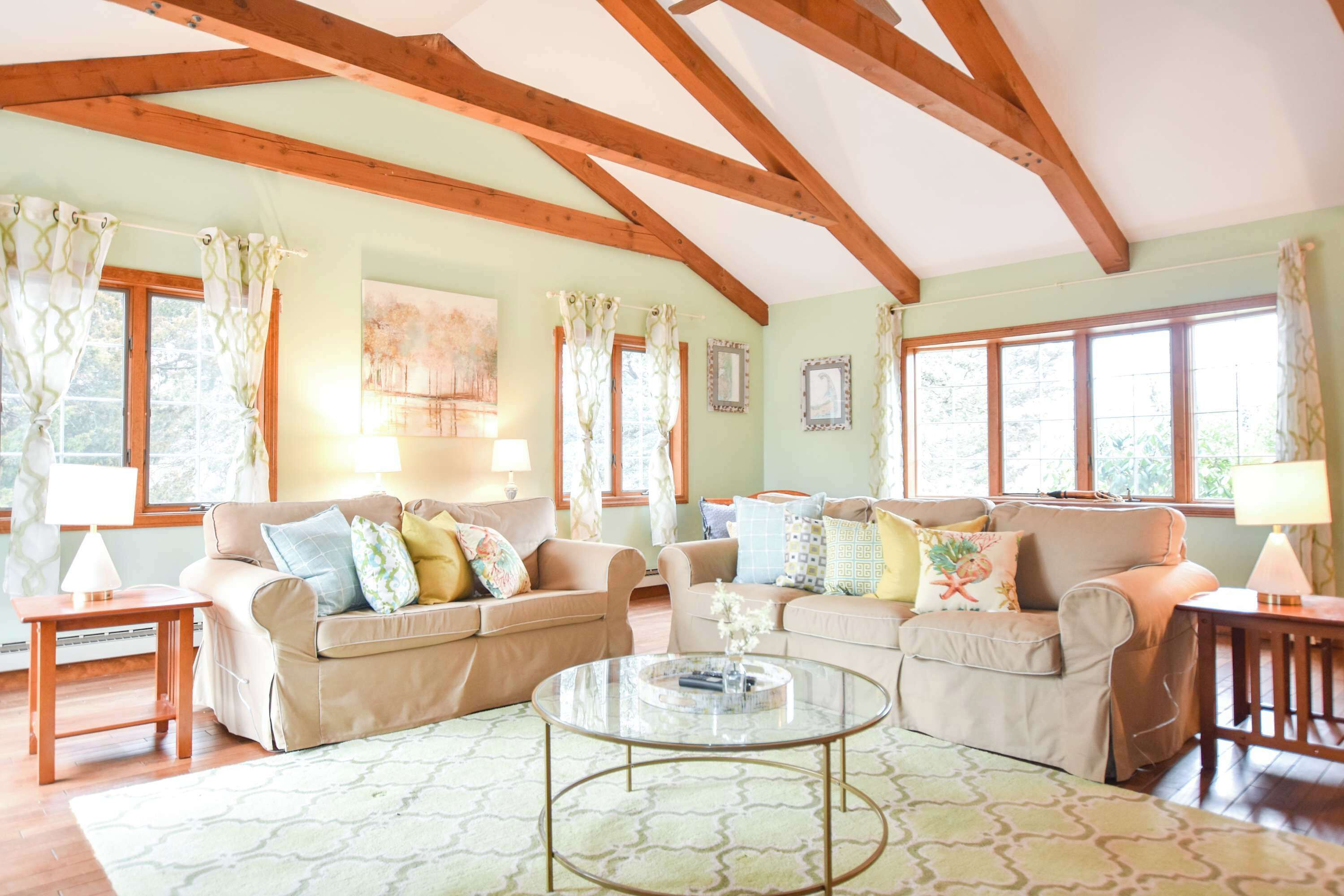 A spacious living room with two sofas, a glass coffee table, wooden beams on the ceiling, and large windows with curtains.