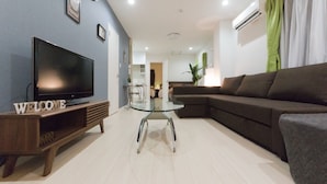 A modern living room with a flat-screen TV on a wooden stand, a glass coffee table, a dark-colored sofa, and a green curtain. - Mini Hotel Shinjuku Front 101 (Tokyo)